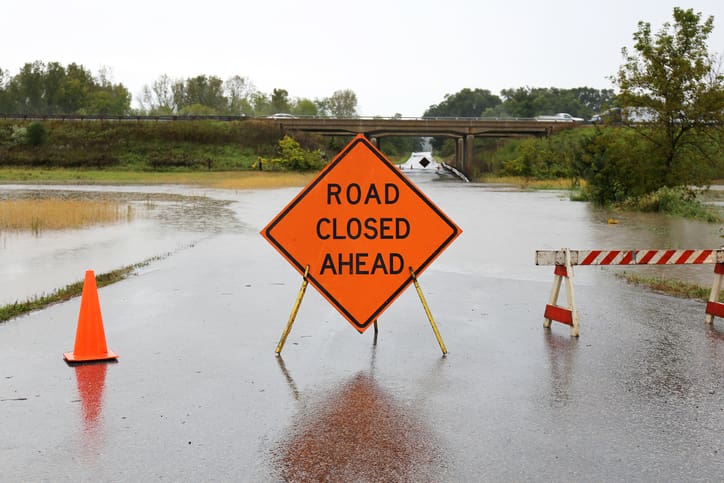 A bright orange Road Closed Ahead Sign and caution cones and barricades are blocking a rain-flooded rural street.