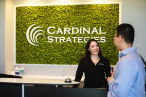 Home page. Brittany and Minh talking in lobby with Cardinal Strategies logo on wall in the background