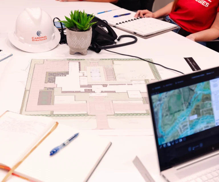 Engineering Services: Cardinal Strategies conference table with a white hard-hat, map, pens, and laptop scattered.