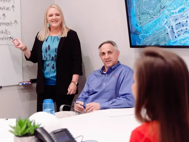 Careers: Image with blonde woman standing at whiteboard, smiling showing something to a man and woman at a conference room table
